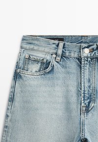 Zone de la taille d'un jean en denim bleu clair avec fermeture par bouton, passants de ceinture, poches avant et détails de couture visibles.