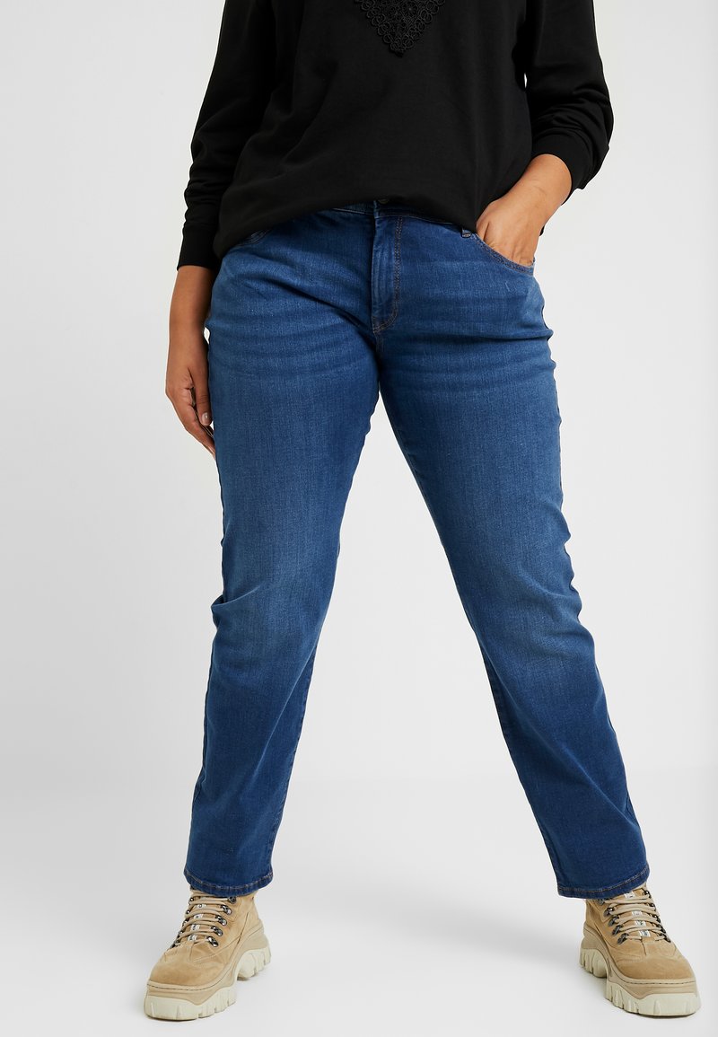 Jeans de mezclilla azul con corte recto, que cuentan con cinco bolsillos, pasadores para cinturón y un ligero desgastado. Combinados con zapatos beige de cordones.