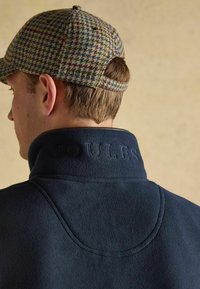 Mann, der eine Kappe mit Hahnentrittmuster und eine marineblaue Fleecejacke mit erhabenem "JOULES"-Logo am hinteren Kragen trägt, vom Kamerawinkel weggewandt.