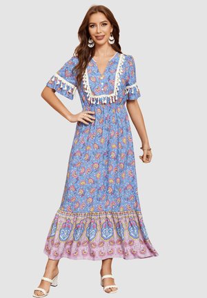 Mujer que lleva un vestido largo azul y rosa con estampado floral y detalles de borlas en las mangas y el escote, combinado con sandalias blancas con tiras trenzadas.