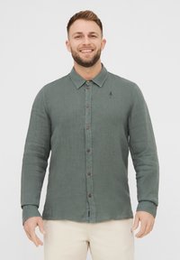 Camicia verde con bottoni, realizzata in tessuto testurizzato, con colletto, bottoni in legno e un piccolo ricamo di un albero sul petto.