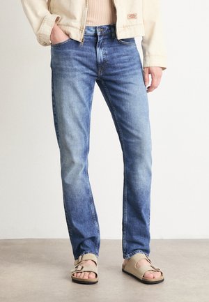 Lichtblauwe denim jeans met een rechte pasvorm, subtiele vervaging en een vijf-pocket ontwerp, gecombineerd met beige sandalen en een crèmekleurige jas.