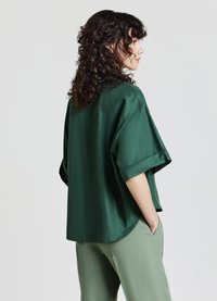 Dark green short-sleeve silk blouse with a relaxed fit, featuring a rounded hem and side slits, paired with light green trousers.