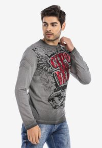 Graues Langarm-T-Shirt mit rot und schwarz gestaltetem Grafikdesign und Flügelmotiven sowie stilisiertem Text. Weicher Stoff mit lockerer Passform.