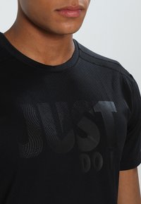 Camiseta deportiva negra con tela texturizada. Presenta una impresión audaz y mate con el texto "JUST DO IT" en el pecho y un escote redondeado.