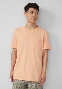 Licht perzikkleurig t-shirt met korte mouwen, gemaakt van zachte stof, met een ronde hals en een borstvak aan de linkerzijde. Textuurafwerking, gecombineerd met gemusterde groene shorts.