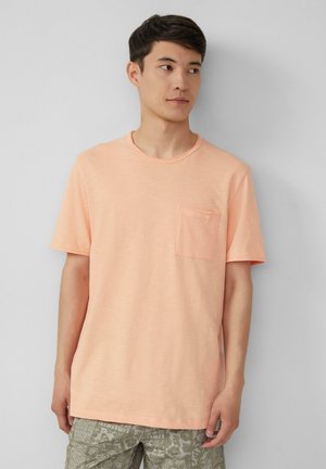 Helles Pfirsichfarbenes T-Shirt mit kurzen Ärmeln aus weichem Stoff, Rundhalsausschnitt und Brusttasche auf der linken Seite. Strukturiertes Finish, kombiniert mit gemusterten grünen Shorts.