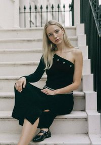 Haut noir asymétrique avec des détails en œillets, associé à une jupe noire côtelée midi avec une fente sur le côté et des ballerines noires avec des accents.