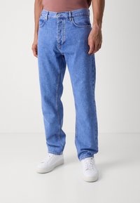 Vaqueros de mezclilla azul claro con corte relajado, con cremallera, cinturilla y un acabado desgastado, combinados con zapatillas blancas.