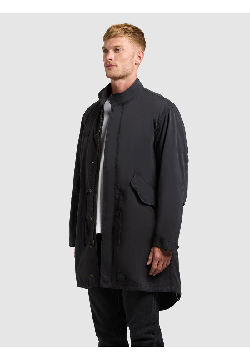 Homme portant un long manteau noir avec des poches à rabat sur une chemise blanche et un pantalon noir, debout devant un fond gris clair uni.
