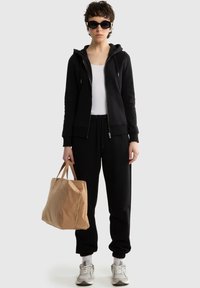 Osoba w czarnej bluzie z kapturem, czarnych spodniach dresowych, białym topie, szarych trampkach, dużych okularach przeciwsłonecznych, trzymająca beżową torbę na tle jednolitego tła.