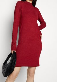 Rode gebreide jurk met lange mouwen, een hoge halslijn en geribbelde manchetten. Gecombineerd met een zwarte gewatteerde handtas. Gladde textuur, getailleerde silhouet.