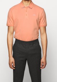 Persikka polo-paita, jossa on kaulus, jossa on valkoiset ja punaiset raidat, kaksivälinen nappisulku ja brodeerattu logo. Yhdistetty tummanharmaisiin muotolaisiin housuihin.