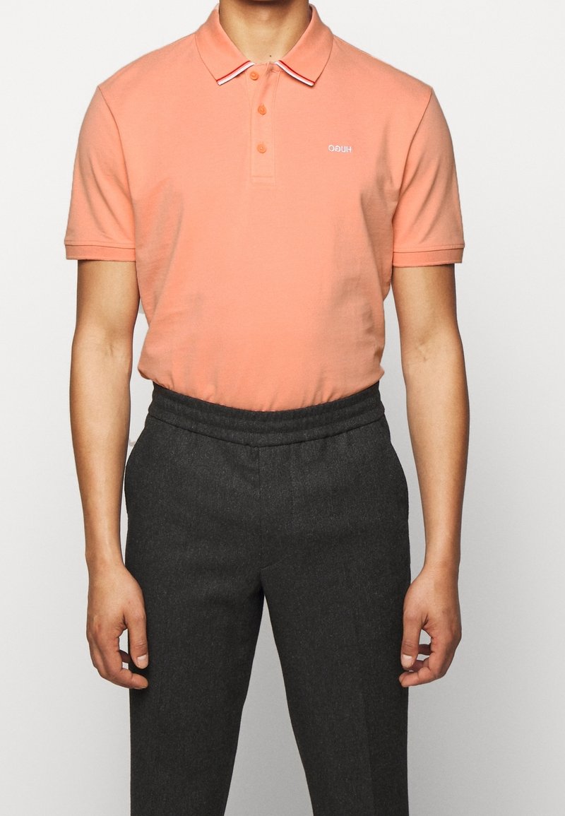 Persikka polo-paita, jossa on kaulus, jossa on valkoiset ja punaiset raidat, kaksivälinen nappisulku ja brodeerattu logo. Yhdistetty tummanharmaisiin muotolaisiin housuihin.