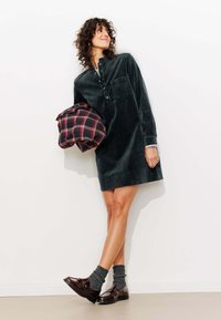 Robe chemise en velours côtelé vert avec un col à volants, une poche avant et une fermeture à boutons. Le modèle tient un sac à carreaux, assorti avec des chaussettes et des chaussures.