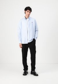 Light blue button-up shirt with a collar, white t-shirt underneath, black trousers, and black shoes; features a small logo on the shirt.