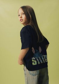 T-shirt noir avec un imprimé holographique "STITCH" et une illustration de personnage bleue au dos, associé à un jean bleu clair.
