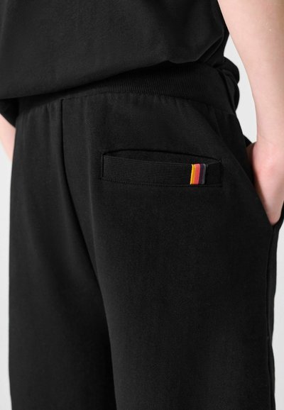 Gros plan sur un pantalon noir avec une poche arrière portant une petite étiquette en tissu aux rayures jaune, rouge et noir. Mains partiellement dans les poches latérales.