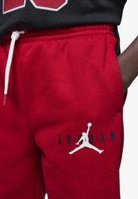 Pantalones de chándal rojos hechos de tela suave con una cintura elástica, que presentan un logo Jumpman en blanco y "JORDAN" impreso en negro.