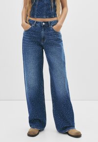 Jeans blu a gamba larga realizzati in denim con borchie bianche sparse, dotati di vita alta e design tradizionale a cinque tasche.