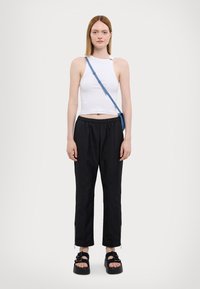 Weißes ärmelloses Oberteil, schwarze Hose mit elastischem Bund, schwarze Plattformsandalen mit Riemen, blaue Umhängetasche, Model steht vor schlichtem Hintergrund.