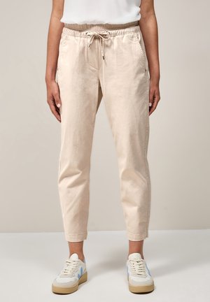 Beige Kordelhose mit Seitentaschen, getragen mit weißen und blauen Sneakern, oberhalb der Knöchel gekürzt, an einer Person, die vor einem schlichten Hintergrund steht.