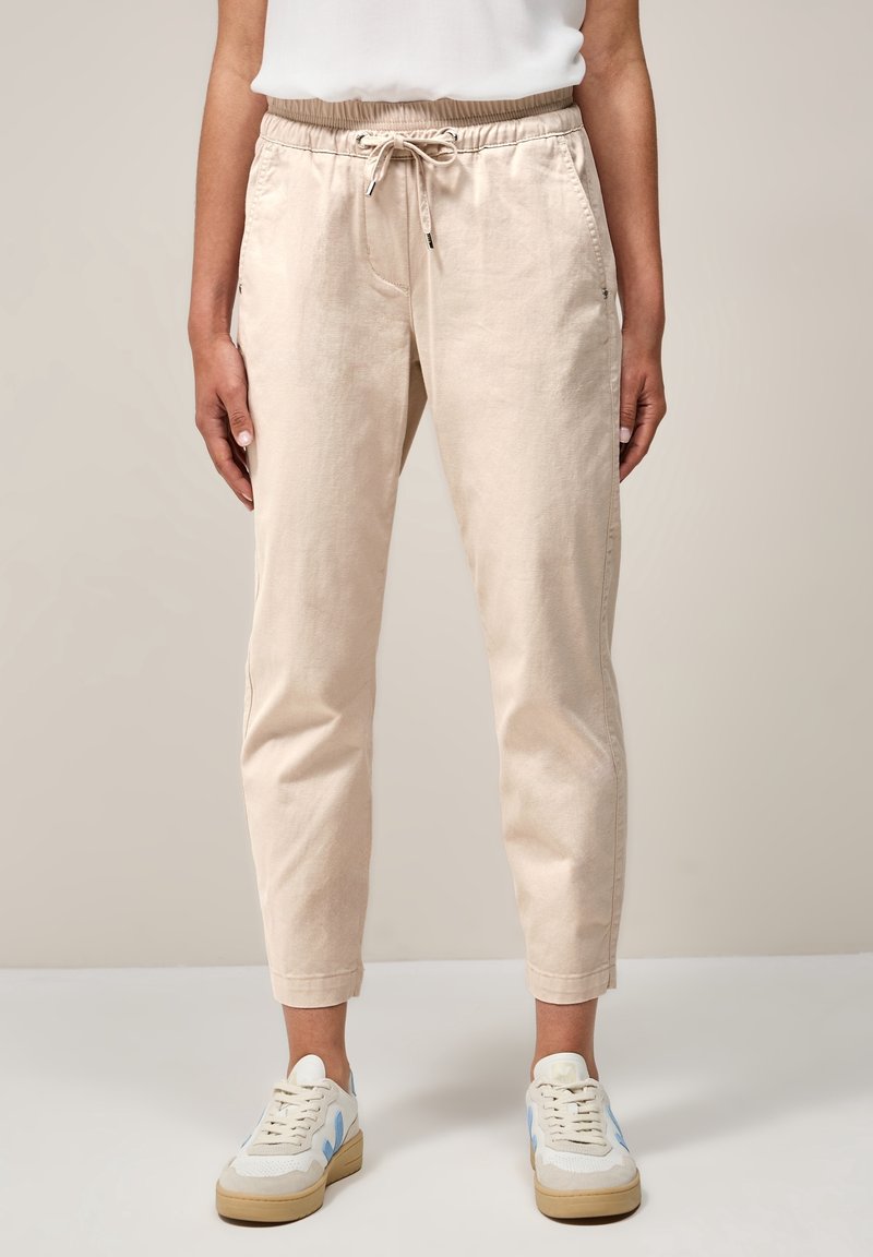 Pantalon beige à cordon avec poches latérales, porté avec des baskets blanches et bleues, coupé au-dessus des chevilles, sur une personne debout devant un fond uni.
