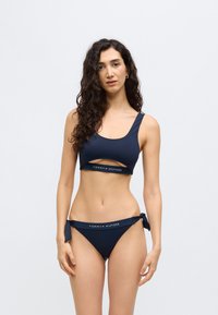 Σετ μπικίνι σε ναυτικό μπλε χρώμα, που περιλαμβάνει crop top με λεπτομέρεια κοψίματος και κάτω μέρος με δεσίματα στο πλάι, κατασκευασμένο από απαλό ελαστικό ύφασμα.