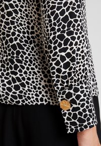 Blouse imprimé animal noir et blanc à manches longues, avec un accent bouton doré texturé sur le poignet et une coupe décontractée.