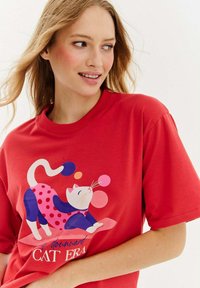Czerwona bawełniana koszulka z krótkim rękawem, na której znajduje się kolorowa grafika kartonowego kota w kropki i zabawne bąbelki. Luźny krój.