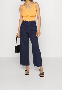 Orange ribbat inknutningsblus med scallopade kanter, ihopparad med högmidjade vida marinblå denimbyxor med framfickor och en knapp.