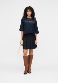 Μπλε ναυτικό μπλουζάκι σε μεγάλο μέγεθος με λευκό λογότυπο "TOMMY HILFIGER", μαύρη πλισέ φούστα, καφέ μπότες μέχρι το γόνατο και λευκή τσάντα χειρός.