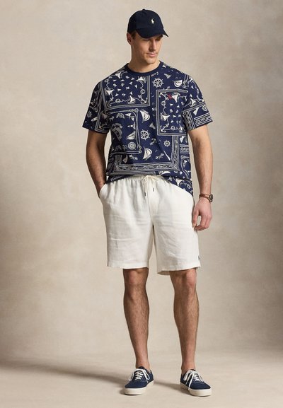Uomo che indossa un berretto blu navy, una t-shirt blu navy con stampa nautica, pantaloncini bianchi con coulisse e sneakers blu navy, in piedi con le mani nelle tasche.