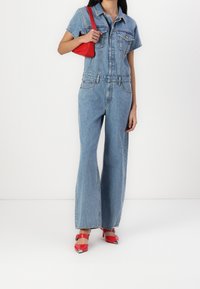 Mono de mezclilla en azul claro, con mangas cortas, bolsillos frontales, cintura entallada y piernas anchas. Combinado con zapatos de tacón rojos y un bolso pequeño rojo.