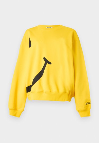 Sweat-shirt jaune à manches longues avec un grand motif abstrait de smiley noir et le logo "Moschino" sur le poignet de la manche gauche.