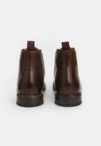 Bocanci din piele maro, până la gleznă, cu un finisaj lucios, având panouri elastice pe lateral și un toc din lemn stratificat. Detaliu de cusătură întărit la cusături.