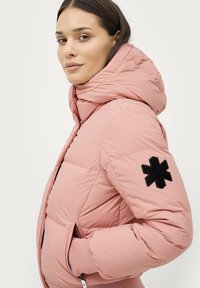 Lyserød pufferjakke med hætte, der har en tekstureret sort detaljeret lap på ærmet og lynlåslommer. Blød, quiltet design.