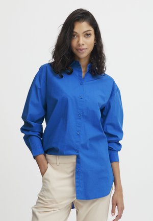 Blue button-up shirt made of a smooth fabric, featuring long sleeves and a collar, paired with cream-colored pants.