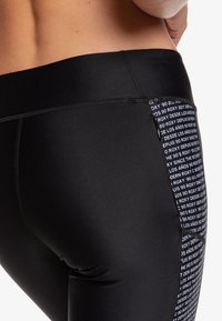 Detailní záběr osoby v černých sportovních legínách s vzorovým postranním panelem, na kterém je bílý text a malá kapsička na zip.