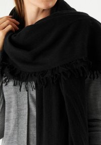 Une écharpe noire en matériau doux avec une texture lisse, dotée d'un bord à franges et drapée sur un pull gris.