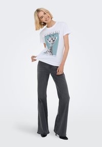 Camiseta de algodón blanca con un gráfico de jirafa sobre un fondo azul verdoso. Combinada con jeans acampanados de color gris oscuro y zapatos de tacón negro.