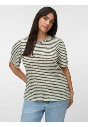 Frau mit langen dunklen Haaren, die ein cremefarbenes Kurzarmshirt mit schwarzen horizontalen Streifen und hellblaue Jeans trägt, vor einem einfarbigen Hintergrund stehend.