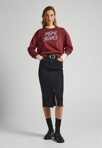 Felpa bordeaux con stampa "PEPE JEANS", indossata sopra una gonna in denim nera aderente con orlo sfrangiato e spacco, abbinata a calze nere e scarpe.