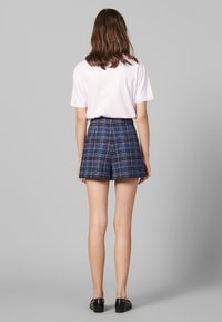 Femme se tenant de dos, portant une chemise à manches courtes rose clair, un short à carreaux bleu taille haute et des mocassins noirs sur un fond gris.