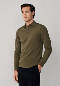 Mladý muž v olivovo zelenom polo tričku s dlhým rukávom a malým červeným logom, v čiernych nohaviciach, stojaci s jednou rukou v vrecku na šedej pozadí.