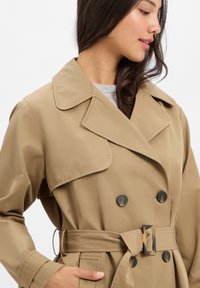Beige Trenchcoat mit einem doppelt brustigen Design, großem Kragen, schwarzen Knöpfen und einem passenden Gürtel. Verfügt über seitliche Taschen und eine glatte Textur.