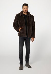 Giacca in pelliccia sintetica marrone con ampio colletto, chiusura con zip e tasche laterali, indossata sopra una maglietta nera, abbinata a jeans scuri e stivaletti grigi.