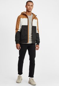Hettejakke med fargeblokker i brun, hvit og svart; glidelås, elastiske mansjetter, kombinert med en grå genser og svarte jeans.