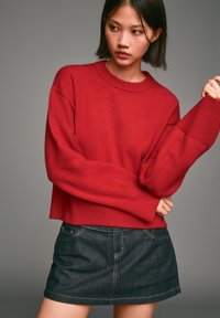 Czerwona sweter wykonany na drutach z okrągłym dekoltem i opuszczonymi ramionami, zestawiony z ciemnoniebieską mini spódnicą z denimu z jasnym stębnowaniem.