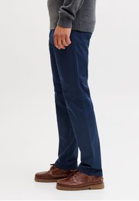 Mand iført marineblå bukser, brune lædermokkasiner og en grå strikket sweater stående mod en hvid baggrund.